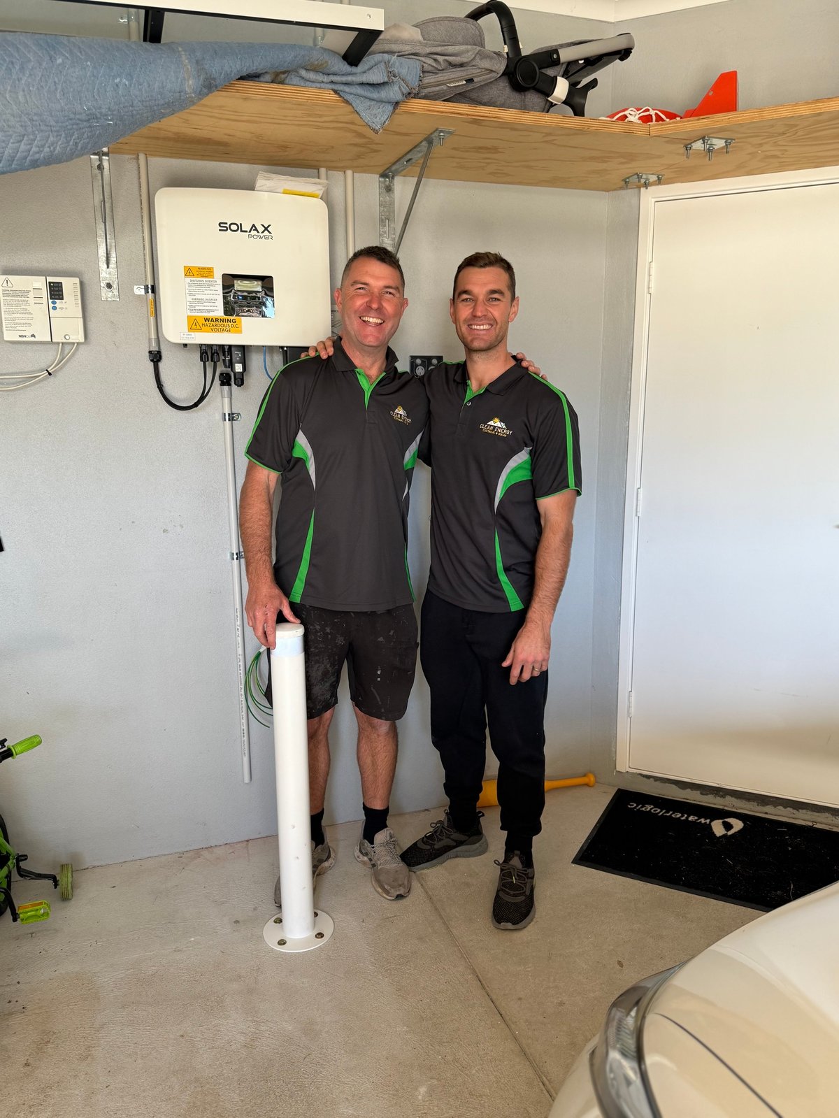 Two men in black and green shirts standing in a garage with solar equipment and white PVC pipe