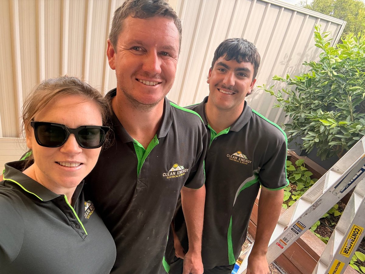 Three people in matching black and green uniforms smiling at camera in greenhouse with plants in background